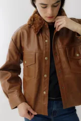 Camisa color marrón de cuero sintético con cuello de piel sintética color marrón claro, cierre frontal con botones dorados, dos bolsillos con solapa en el pecho y corte recto.