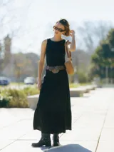 Vestido largo negro sin mangas, con cuello redondo y corte regular. Incluye cinturón marrón con detalles metálicos y botas de cuero negro con hebillas.