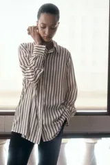 Camisa de crepé color crema con rayas verticales marrones, corte oversized, cuello clásico, cierre frontal con botones ocultos, mangas largas con puños abotonados, pliegue de caja invertido en la espalda y bajo recto.