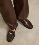 Zapato mocasín de cuero color marrón oscuro con detalle metálico dorado en la parte delantera.