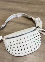 Riñonera blanca de cuero sintético con textura, decorada con tachas metálicas redondas plateadas en el frente. Posee cierre metálico plateado y correa ajustable con tejido a rayas en tonos beige y blanco.