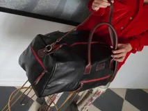 Bolso de viaje tipo weekender de cuero color marrón con detalles en rojo en los cierres. Tiene doble asa corta y correa de hombro ajustable y extraíble. Cuenta con bolsillos internos y externos.