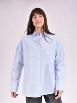 Camisa celeste de corte amplio, con cuello clásico, cierre frontal con botones, bolsillo en el pecho y mangas largas con detalle de botones en los puños.