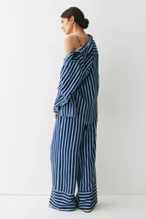 Camisa oversized de sarga a rayas verticales azules y blancas, con cuello de pico y botonadura frontal. Modelo con hombros caídos, mangas largas con tapeta y puños abotonados, y canesú en la espalda. Bajo recto con aberturas laterales redondeadas.