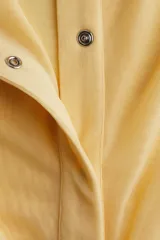 Camisa de malla color amarillo pálido, de corte entallado, con cuello clásico, mangas largas con puños abotonados y cierre frontal con botones a presión ocultos. Presenta frunces en la parte delantera que crean un efecto drapeado.