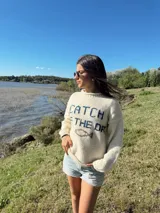 Sweater color crema tejido en lana merino, con cuello redondo y mangas largas. Presenta la frase "CATCH THE DAY" en color azul y un dibujo de un pez en el centro.
