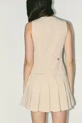 Vestido corto color beige, de escote pico y sin mangas. Tiene falsos bolsillos de vivo en el delantero, falda con tablas y detalle de trabilla en la espalda. Cierre lateral con cremallera oculta.