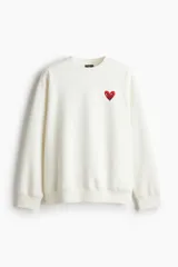 Buzo blanco de corte holgado con cuello redondo y puños y bajo acanalados. Presenta un pequeño logo de corazón rojo con ojos en el pecho.