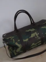 Bolso tipo duffel de lona con estampado de camuflaje y detalles en cuero marrón. Tiene doble asa de mano y correa de hombro ajustable y extraíble.