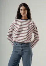 Camiseta de manga larga, corte holgado, con rayas horizontales rojas y blancas. Posee cuello redondo con ribete azul marino y un pequeño logo bordado en el pecho.