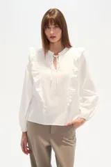 Blusa blanca de calce holgado con cuello en V y lazo frontal. Presenta detalles de volados en el pecho y cuello, y mangas largas con puños elásticos.