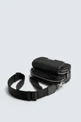 Bandolera negra de nylon con correa ajustable y removible. Tiene un compartimento principal con cierre de cremallera, un bolsillo interior plano con cremallera y un bolsillo frontal con solapa y cremallera.