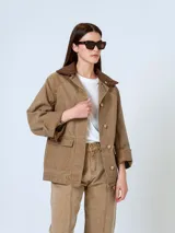 Chaqueta color marrón claro con cuello de pana marrón, cierre con botones a presión, mangas largas con puños abotonados, bolsillos delanteros tipo parche y corte recto.