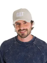 Gorro de béisbol color beige claro, confeccionado en sarga de algodón teñida con pigmentos. Presenta visera curva y el logo de Caterpillar (CAT) bordado en blanco en el panel frontal. Posee ajuste trasero con broche metálico.