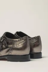 Zapato bajo de cuero negro, estilo monk strap con dos hebillas ajustables sobre el empeine. Presenta detalles de brogue (perforaciones) en la puntera y alrededor del borde superior. Tiene un taco bajo y ancho.