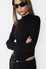 Body manga larga color beige, de punto roma, con cuello medio alto con cierre de nylon a tono. Tiene recortes pisados con bordador, mangas ranglan con mitones en los puños, broches snap en la entrepierna y elástico interno en el contorno de tiro.