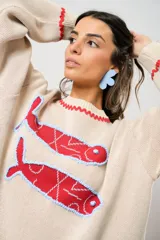 Sweater tejido color crudo con cuello redondo y mangas largas. Presenta dos peces rojos con detalles en azul bordados en el frente. Tiene ribete ondulado rojo en cuello, puños y ruedo.