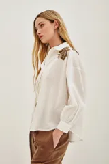 Camisa blanca de algodón con cuello clásico, mangas largas abullonadas con puños anchos y cierre frontal de botones. Presenta aplicaciones bordadas en dorado con detalles de animal print en los hombros.