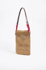 Bolso tote de rafia color beige con diseño calado, asas cortas de cuero marrón y cordones rojos decorativos.