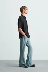 Camisa de hombre de manga corta, cuello solapa y corte relaxed fit. Tejido blanco con textura sutilmente arrugada o plisada. Cierre frontal de botonadura y bajo con aberturas laterales.