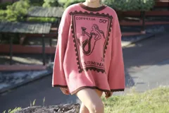 Buzo oversized de punto color tostado con diseño gráfico frontal en azul inspirado en una estampilla con una sirena y el texto 'Taormina'. Presenta tajos laterales, cuello redondo y terminaciones en contraste de color azul.