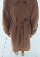 Abrigo largo color marrón camel, confeccionado en mezcla de lana. Presenta cuello con solapas, cierre frontal con botones, mangas largas con botones en los puños, bolsillos laterales y cinturón a tono.