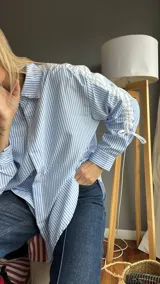 Camisa celeste a rayas verticales blancas, de corte holgado, con cuello clásico, mangas largas con ajuste de lazos y detalle de volados en los hombros.