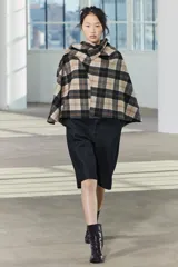 Capa de cuello alto confeccionada en tejido con lana a cuadros en tonos beige, negro y marrón. Tiene bajo asimétrico y cierre en la espalda con botón.