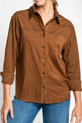 Camisa marrón de gamuza vegana con cuello clásico, cierre frontal con botones y mangas largas con puños abotonados. Presenta detalles de tachas metálicas en cuello y charreteras.