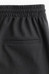 Pantalón negro de sarga suave con cintura elástica, cordón de ajuste en color contrastante y franjas laterales finas. Modelo de corte holgado con tiro alto, bolsillos laterales discretos y bolsillos insertados decorativos atrás. Piernas amplias con pliegues adelante y atrás.