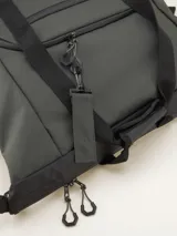 Bolso tipo duffel verde militar con base y detalles en negro. Tiene doble asa de mano y correa de hombro ajustable y extraíble. Cuenta con dos bolsillos frontales con cierre y logo de la marca en la parte inferior.