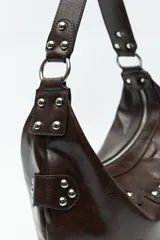 Cartera marrón de cuero sintético con forma de media luna, asa corta y apliques de tachas metálicas plateadas.