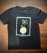 Remera negra de corte clásico con estampado en la espalda de un paisaje de atardecer con palmeras en un marco rectangular.