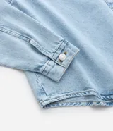 Camisa de jean de algodón, color celeste claro lavado, con cuello tradicional, manga larga y bolsillo de parche en el pecho. Presenta un cierre frontal con botones metálicos.