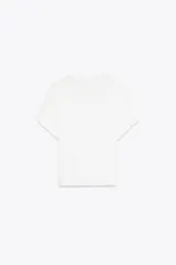 Camiseta blanca de algodón con cuello redondo y manga corta. Presenta bordados florales a tono en el lateral derecho del pecho y en la parte inferior izquierda.