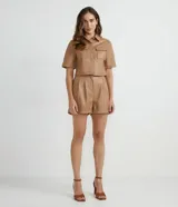 Camisa de manga corta confeccionada en material sintético color beige. Tiene cuello camisero, cierre de botones en forma de corazón, dos bolsillos con solapa y corte cropped.