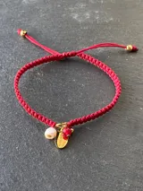 Pulsera tejida con hilo rojo y dijes colgantes: medallita dorada con mensaje Good Luck, perlita de río, perla de acero dorada y cuenta de jade roja.