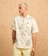 Camisa de manga corta con estampado de palmeras y motivos playeros en tonos celeste y verde sobre fondo blanco.