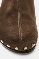 Zueco destalonado de piel color marrón con acabado en punta redonda. Presenta detalle de tachas doradas en los laterales y pespunte decorativo en la parte delantera. Posee una suela plana con un tacón mínimo de 0,5 cm.