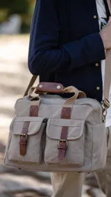 Bolso tipo duffel de lona color beige con detalles en cuero marrón. Tiene doble asa de mano y correa bandolera ajustable y desmontable. Cuenta con cierre superior, bolsillos frontales con hebilla y bolsillos internos.