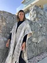 Ruana larga de lana merino color beige jaspeado, con diseño de trenzas y flecos en el borde inferior.