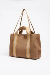 Bolso shopper grande de estructura blanda, color marrón, confeccionado en nylon con detalles de piel vacuna. Presenta doble asa de mano con cinta de tejido beige y naranja, y un logo metálico de Bimba y Lola en el frente.