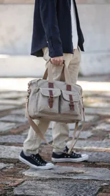 Bolso tipo duffel de lona color beige con detalles en cuero marrón. Tiene doble asa de mano y correa bandolera ajustable y desmontable. Cuenta con cierre superior, bolsillos frontales con hebilla y bolsillos internos.