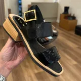 Sandalias negras con dos tiras anchas, una sobre los dedos y otra al tobillo con hebilla dorada. Taco cuadrado de madera clara.