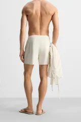 Bañador corto color blanco hueso de tejido técnico, con cintura elástica ajustable con cordón, bolsillos laterales y trasero.