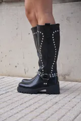 Bota alta de caña ancha color negro, estilo biker o motera, con suela track gruesa y taco bajo. Presenta apliques de tachas plateadas a lo largo de la caña y una correa con hebilla y argolla decorativa alrededor del tobillo.