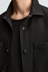 Sobrecamisa negra de tejido con elasticidad, cuello solapa y manga corta. Tiene dos bolsillos de plastrón en el pecho y cierre frontal con botones a presión.