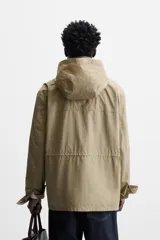 Parka color beige de tejido técnico ligero, corte relaxed fit, cuello alto con capucha plegable, manga larga con puños ajustables con botón a presión, cierre frontal con cremallera, cintura ajustable con elástico interno y múltiples bolsillos frontales de parche con solapa.