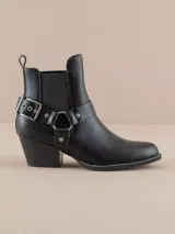 Botín negro de cuero sintético con elásticos laterales, taco bajo y arnés decorativo con hebilla y argolla metálica.