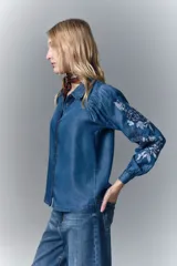 Camisa de calce recto confeccionada en tencel azul denim, con manga larga amplia con frunces en los hombros y cuello camisero. Presenta bordado de hilo en mangas y cartera. Mangas terminadas en puño con botón y ojal. Cartera delantera con botones metálicos con el logo de la marca.